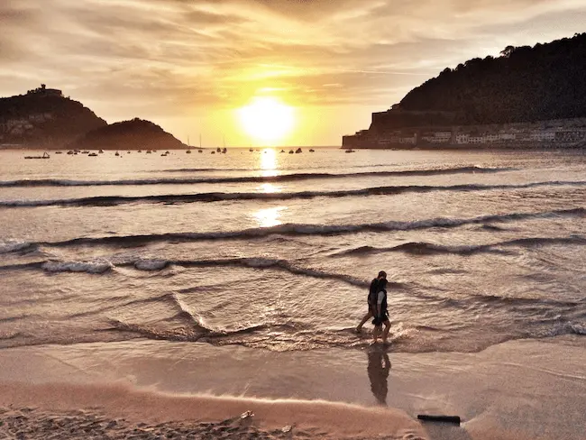 Anochecer en la Playa de la Concha, con una persona paseando a lo largo de la orilla mientras el cielo se tiñe de colores cálidos y las aguas reflejan la luz del atardecer.