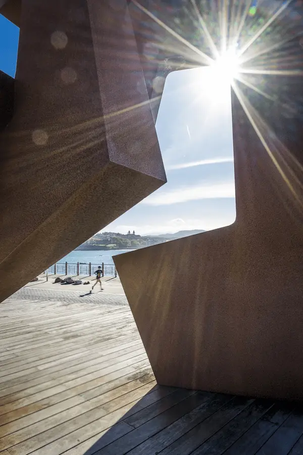 Un hombre camina por un muelle de madera mientras el sol brilla a través de una gran escultura.