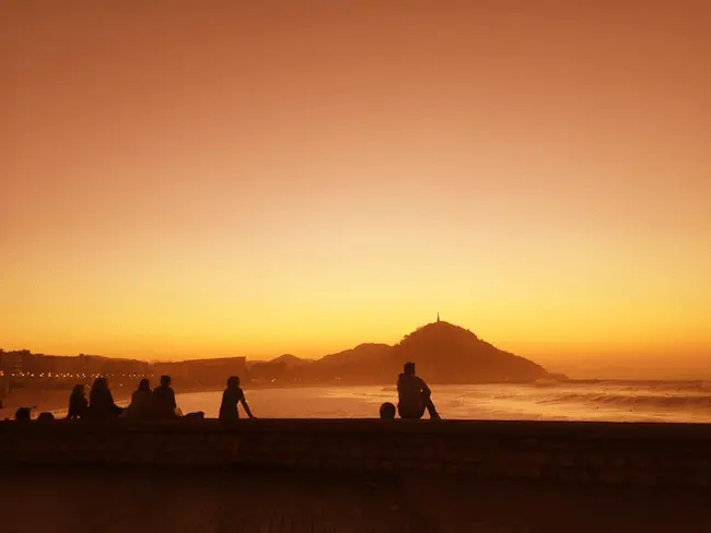 Vista del atardecer desde el muro de Sagües, con siluetas de personas sentadas disfrutando del paisaje, mientras el sol tiñe de naranja el horizonte sobre la playa.