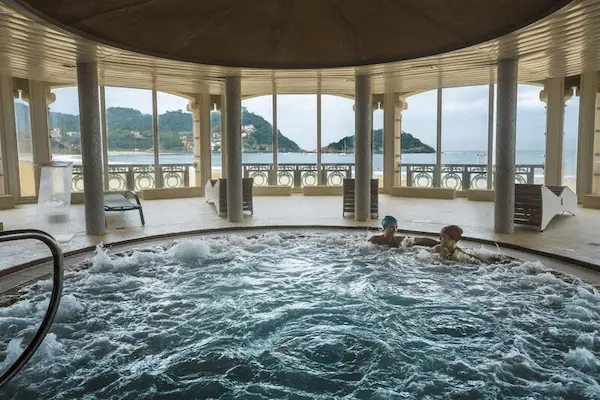 Gran piscina circular con dos personas disfrutando, rodeada de un hermoso paisaje de playa y océano al fondo.