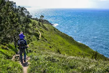 Hombre con niño en hombros por un camino de trekking con vistas al mar