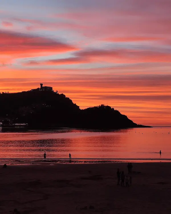 Un cielo rojo se extiende sobre la playa, donde varias personas disfrutan de un paseo al atardecer.