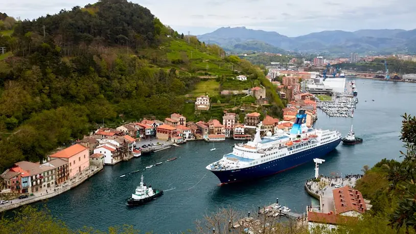 Un gran crucero en el puerto de Pasaia, con casas del pueblo y montañas de fondo.