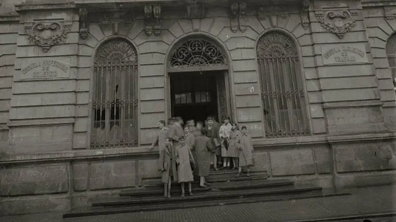 Grupo de alumnos saliendo de la Escuela de Artes y Oficios, posando frente al edificio en un retrato antiguo.