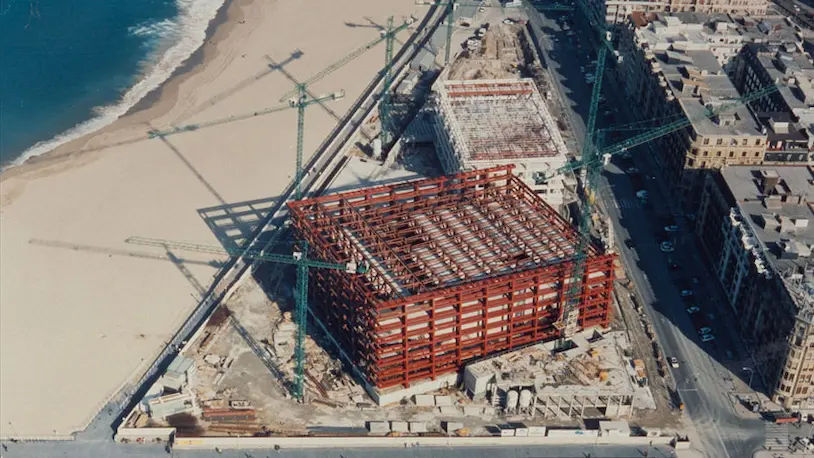 Vista aérea del Edificio Kursaal en construcción, ubicado junto a la playa, con grúas y materiales de construcción visibles.
