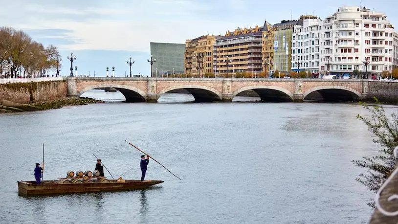 Tres hombres reman en un bote sobre el río Urumea, con el puente de María Cristina al fondo.