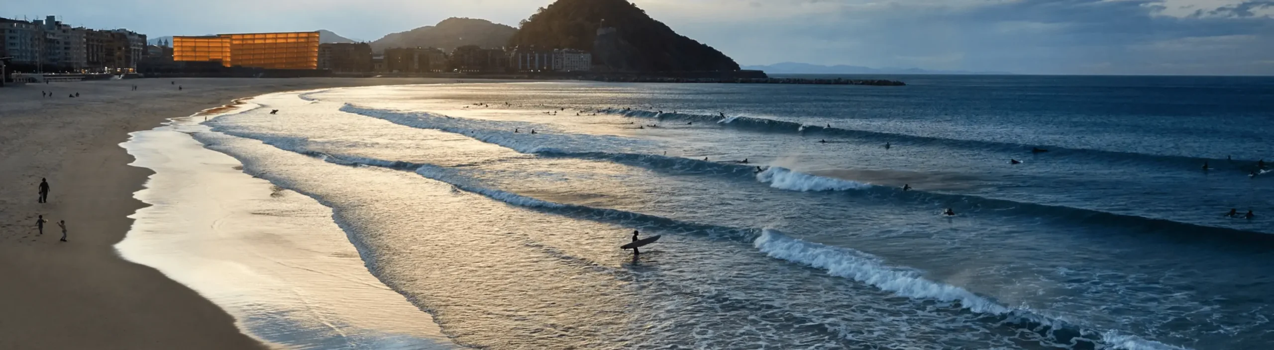 Panorámica de la playa al atardecer con surfistas y el Kursaal iluminado