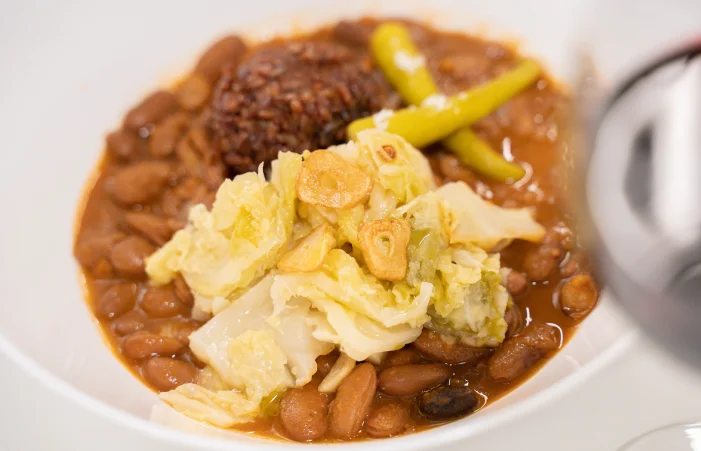 Un primer plano de un plato tradicional vasco de alubias rojas (tolosanas o pintas) con sus sacramentos: berza cocida con ajos fritos, arroz oscuro en el centro y guindillas de Ibarra. El plato está servido en un bol blanco y la imagen tiene un ligero desenfoque en la parte derecha donde se aprecia el borde de un recipiente.