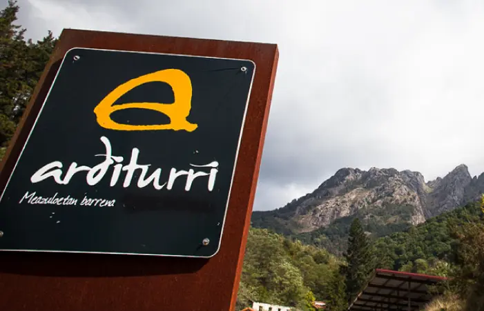 Primer plano de un cartel de "Arditurri" con montañas y un cielo nublado al fondo.