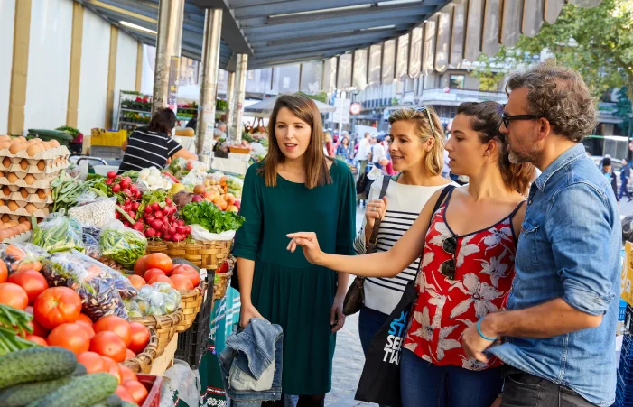 Un grupo de cuatro personas mirando y seleccionando productos frescos en un puesto de un mercado al aire libre.