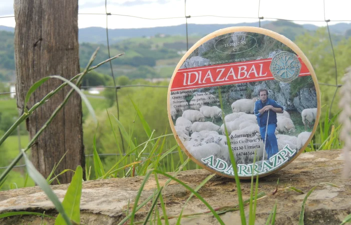 Una rueda entera de queso Idiazabal "Adarrazpi" con la etiqueta visible, apoyada sobre un muro de piedra en un paisaje rural vasco verde y montañoso.