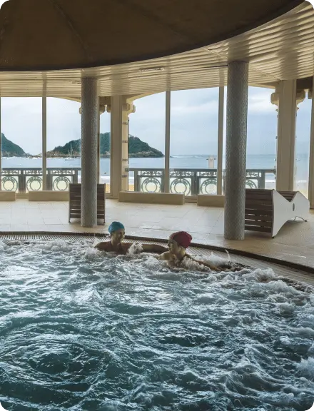 Gran piscina circular con dos personas disfrutando, rodeada de un hermoso paisaje de playa y océano al fondo.