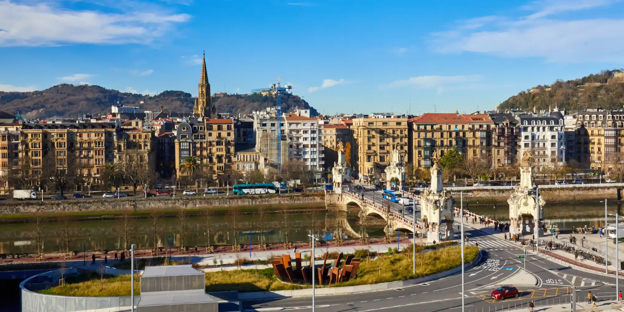 Una vista aérea de la ciudad de Donostia-San Sebastián en un día soleado, mostrando el río Urumea cruzado por el ornamental Puente de María Cristina con sus obeliscos, edificios históricos a lo largo de la orilla, la aguja de la Catedral del Buen Pastor y montañas al fondo.