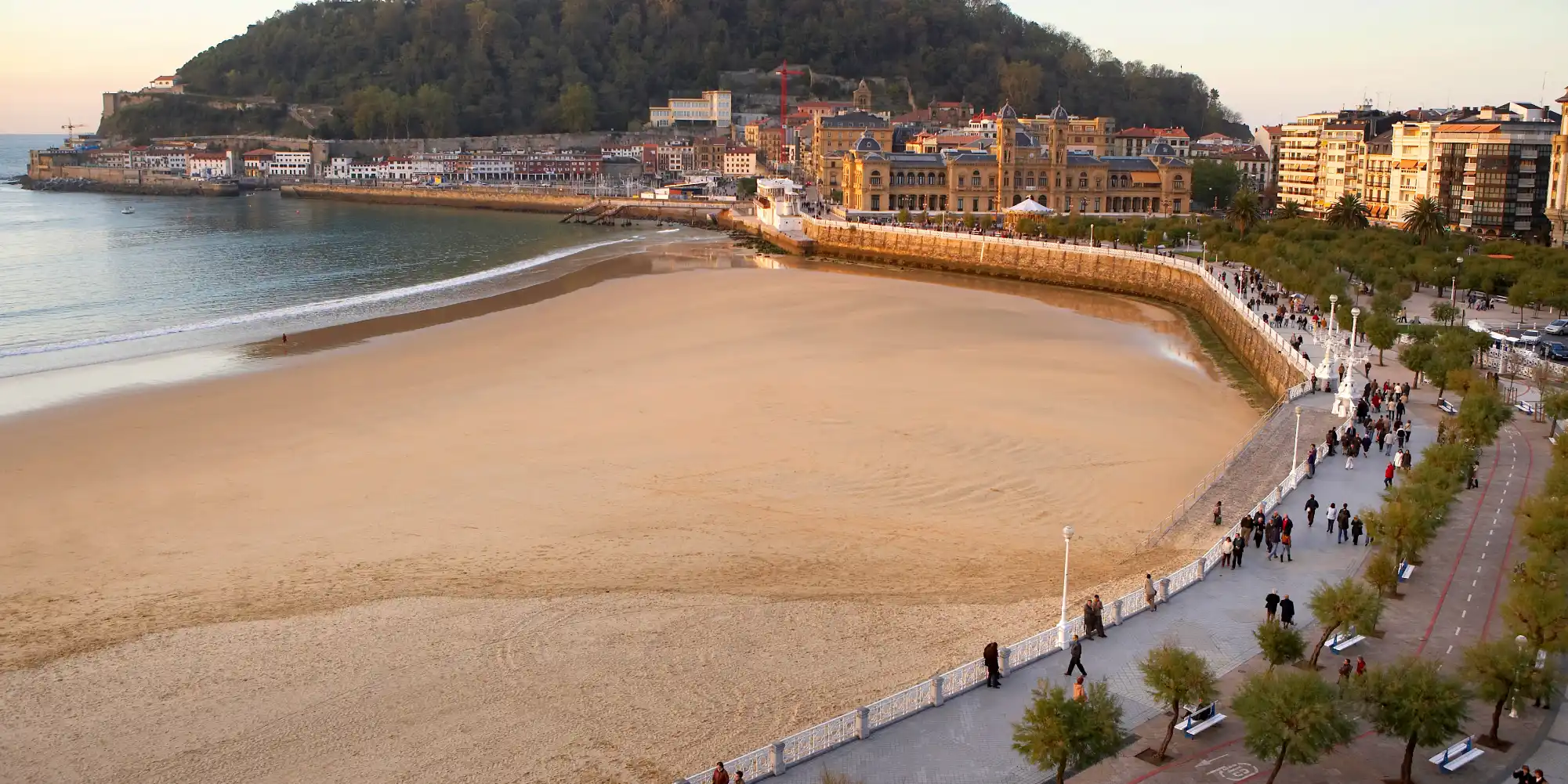Vista aérea de la Bahía de La Concha en Donostia-San Sebastián al atardecer, mostrando la amplia playa de arena dorada, el paseo marítimo curvado con gente paseando, y los edificios históricos de la ciudad con el Monte Urgull al fondo.