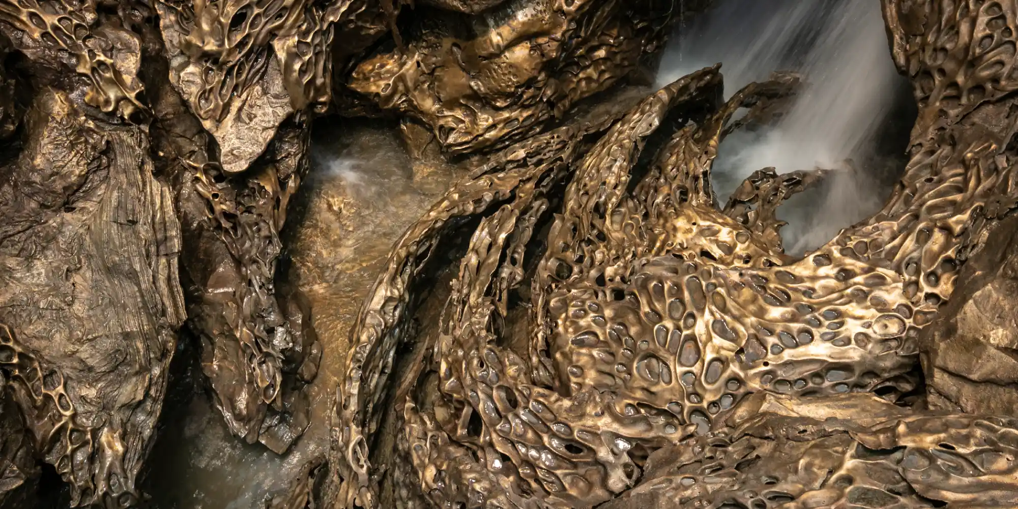 Primer plano de una pared de cueva con formaciones rocosas irregulares y texturizadas de tonos marrones y ocres, parecidas a las caprichosas formas de "macarrones" o "gours", con agua que fluye y cae en una pequeña cascada a la derecha.