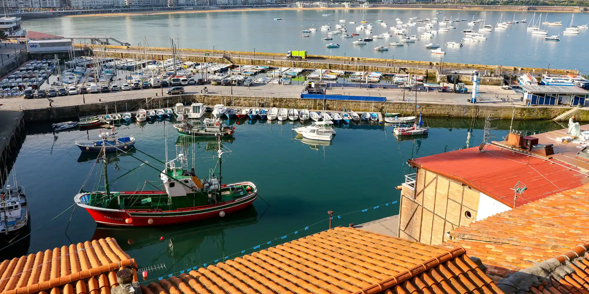 Vista aérea del puerto de Donostia-San Sebastián, con un primer plano de tejados de tejas naranjas y un muelle lleno de embarcaciones de recreo y pesqueras, incluyendo un barco rojo y verde en el agua tranquila, y la Bahía de La Concha con más barcos al fondo.