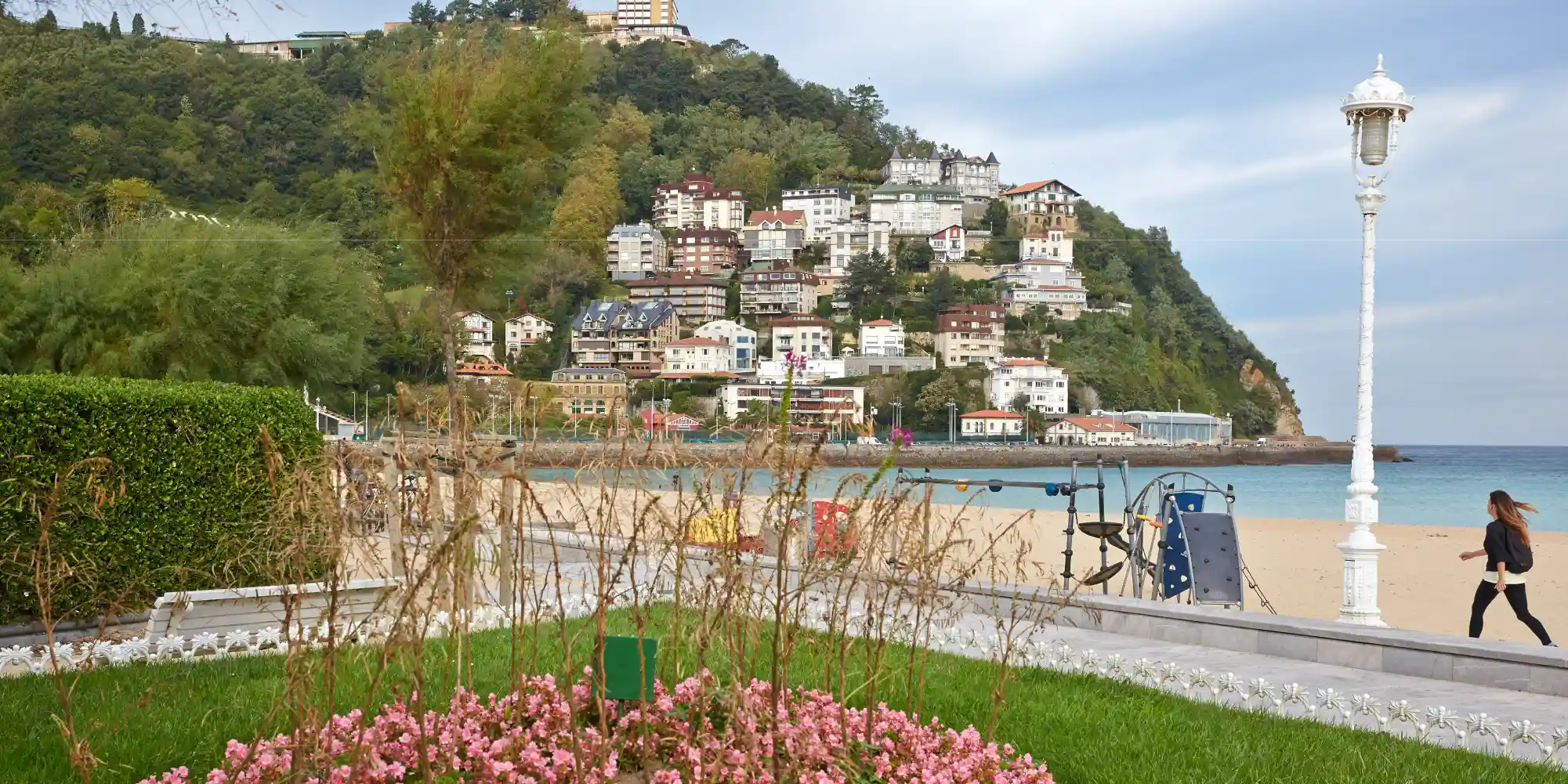 Una vista panorámica de la playa de La Concha en San Sebastián. En primer plano, hay un césped verde con flores rosas y un seto, y a la derecha, un farol blanco ornamentado. Más allá, se extiende una playa de arena dorada con el mar azul claro. Al fondo, una ladera verde cubierta de árboles y edificios de estilo tradicional, culminando en la cima con un gran edificio y el monte Igueldo. Una persona camina por el paseo junto a la playa en el lado derecho de la imagen.