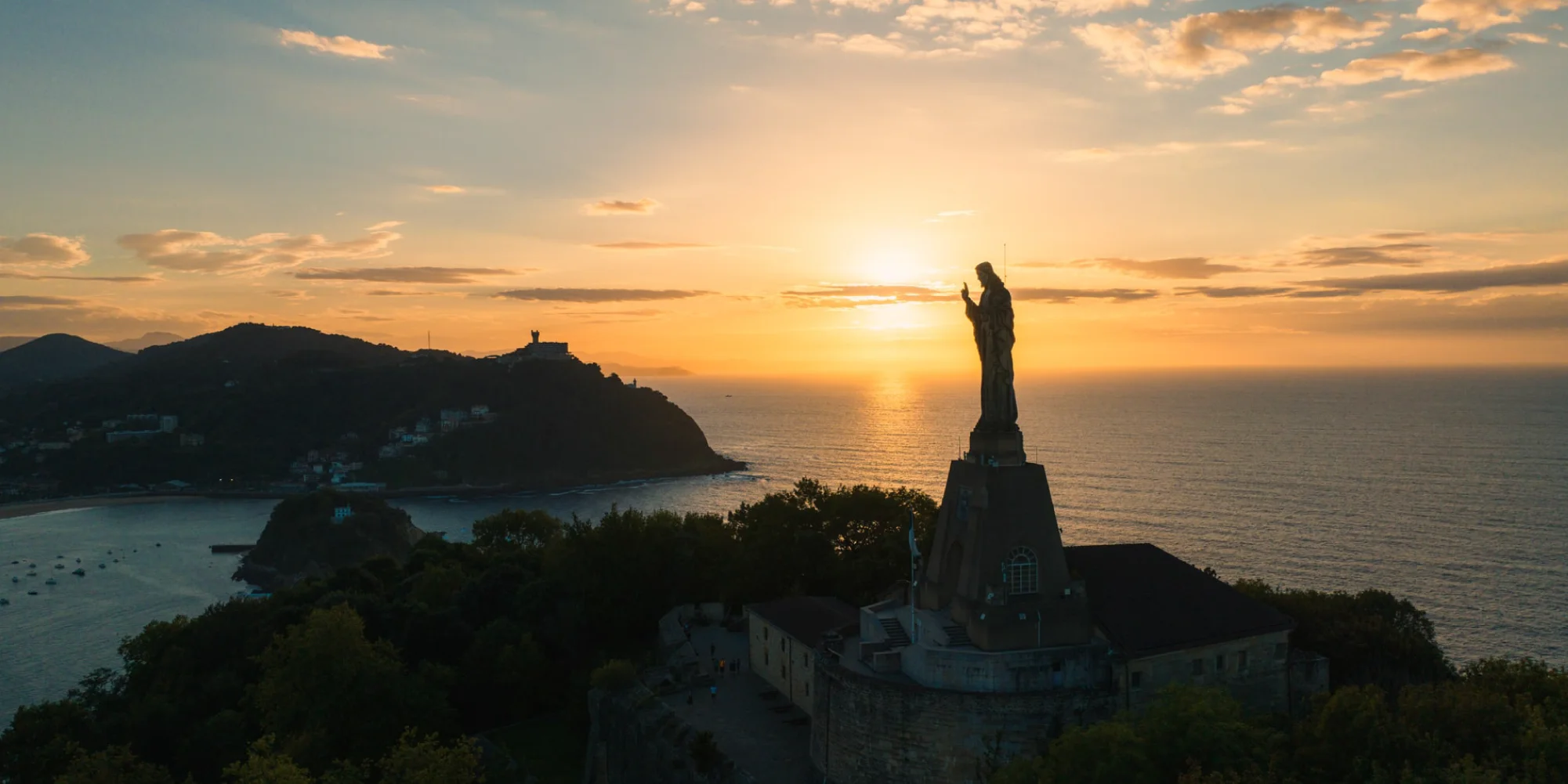 Vista aérea de San Sebastián al atardecer, mostrando la estatua del Sagrado Corazón de Jesús en el Monte Urgull en primer plano. El sol poniente crea un resplandor dorado sobre el horizonte del mar Cantábrico, iluminando el cielo con tonos anaranjados y amarillos. Al fondo, se distingue la bahía de La Concha con barcos anclados y la silueta del Monte Igueldo. La estatua se erige majestuosamente, con su figura recortada contra el cielo brillante.