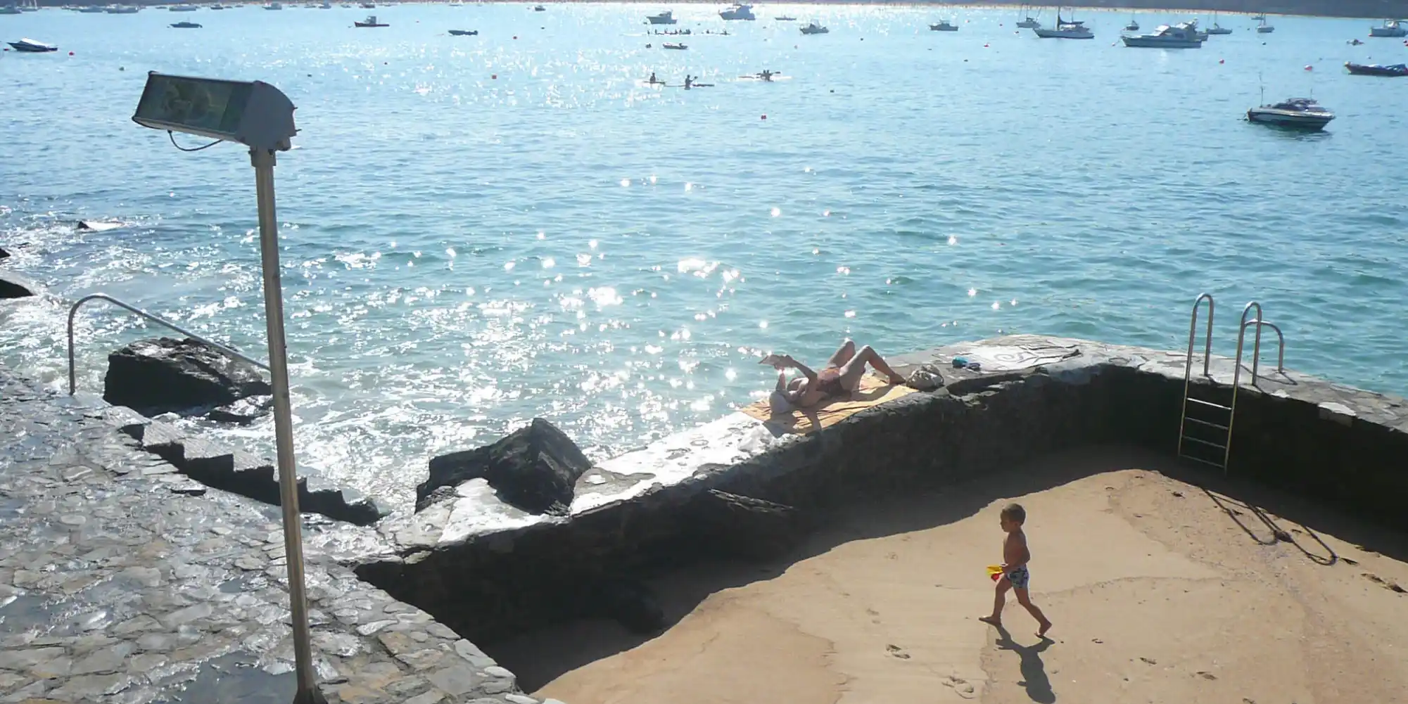 Vista de una playa con arena y acceso al mar, donde un adulto toma el sol y un niño pequeño camina por la orilla, con varias embarcaciones y personas nadando en la distancia, bajo un cielo despejado.