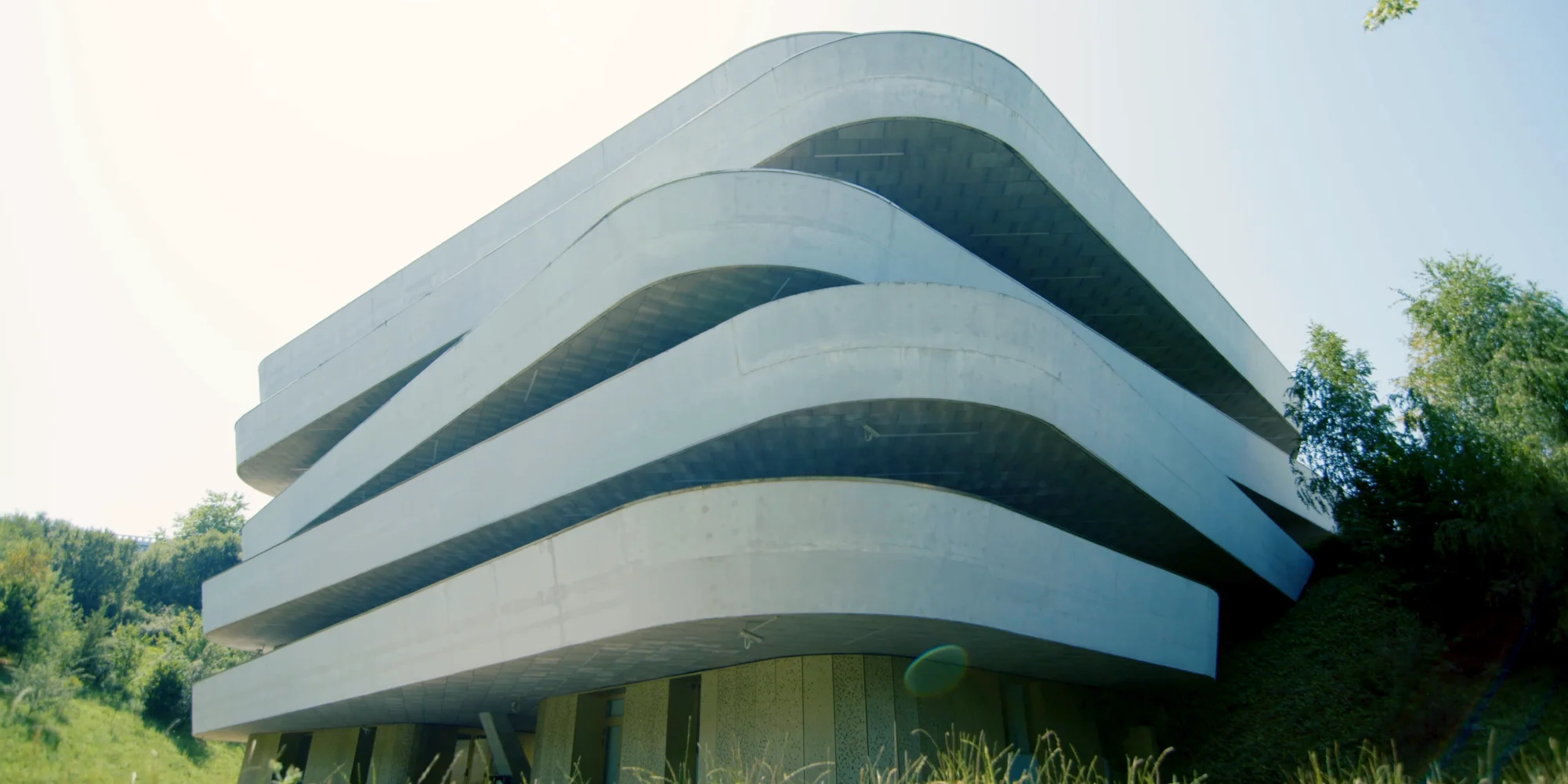 Edificio moderno con capas curvas y horizontales de color claro, rodeado de vegetación bajo un cielo luminoso.