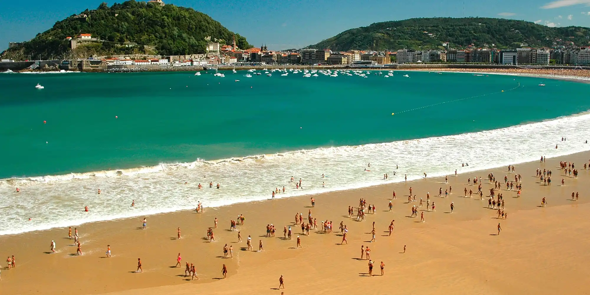 Una vista panorámica de la Bahía de La Concha en Donostia-San Sebastián, con una playa de arena dorada llena de bañistas y gente paseando, el mar azul-turquesa con olas y barcos, y la ciudad con sus montañas verdes al fondo bajo un cielo despejado.