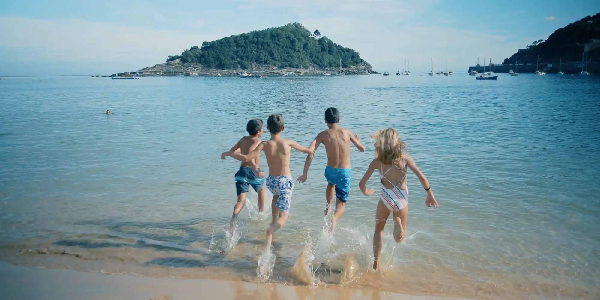 Cuatro niños, de espaldas, corren y salpican en la orilla de una playa de arena dorada, adentrándose en el mar azul. Al fondo se ve una isla montañosa con vegetación, numerosos barcos anclados y la costa de Donostia-San Sebastián.