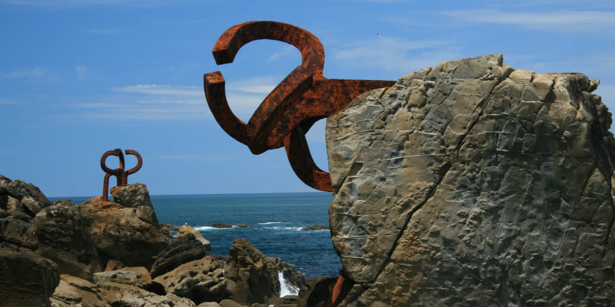 Primer plano de una de las esculturas de acero oxidado del "Peine del Viento" de Eduardo Chillida, encajada en una gran roca con textura. Al fondo, otra escultura similar, el mar Cantábrico con pequeñas olas y el cielo azul, en Donostia-San Sebastián.