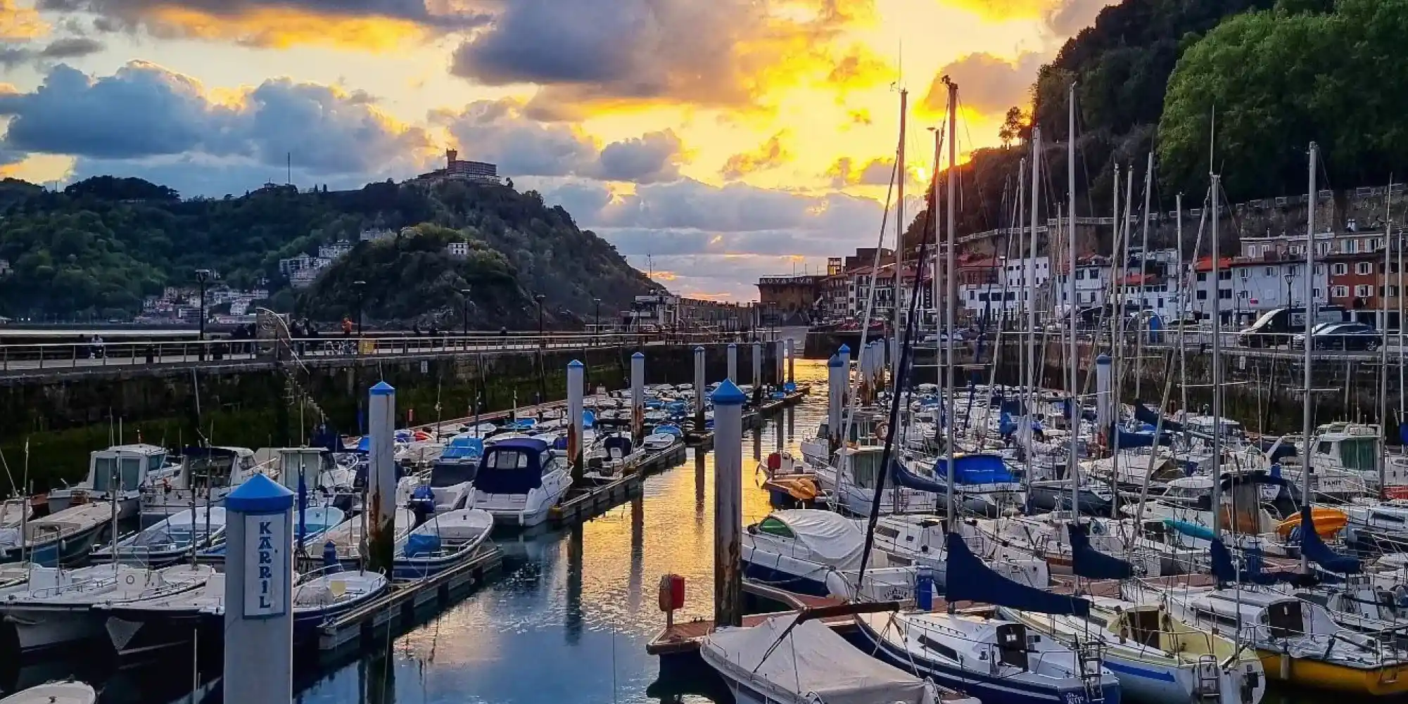 Una marina llena de veleros y embarcaciones de recreo amarradas en el puerto de Donostia-San Sebastián al atardecer, con el cielo teñido de tonos anaranjados y dorados que se reflejan en el agua. Montañas y edificios costeros se vislumbran al fondo.