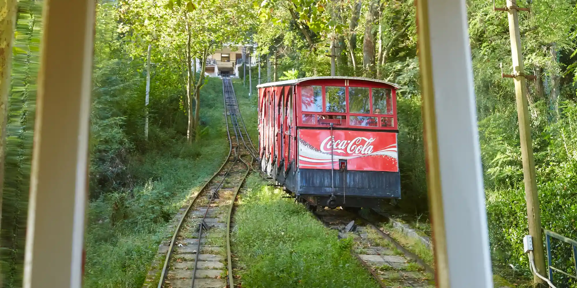 Un funicular rojo brillante con el logotipo de Coca-Cola sube por una vía a través de una densa vegetación verde. Se ve una casa en la cima de la colina al final de las vías, y el tren está enmarcado por lo que parecen ser las ventanas de otro vagón o estructura.
