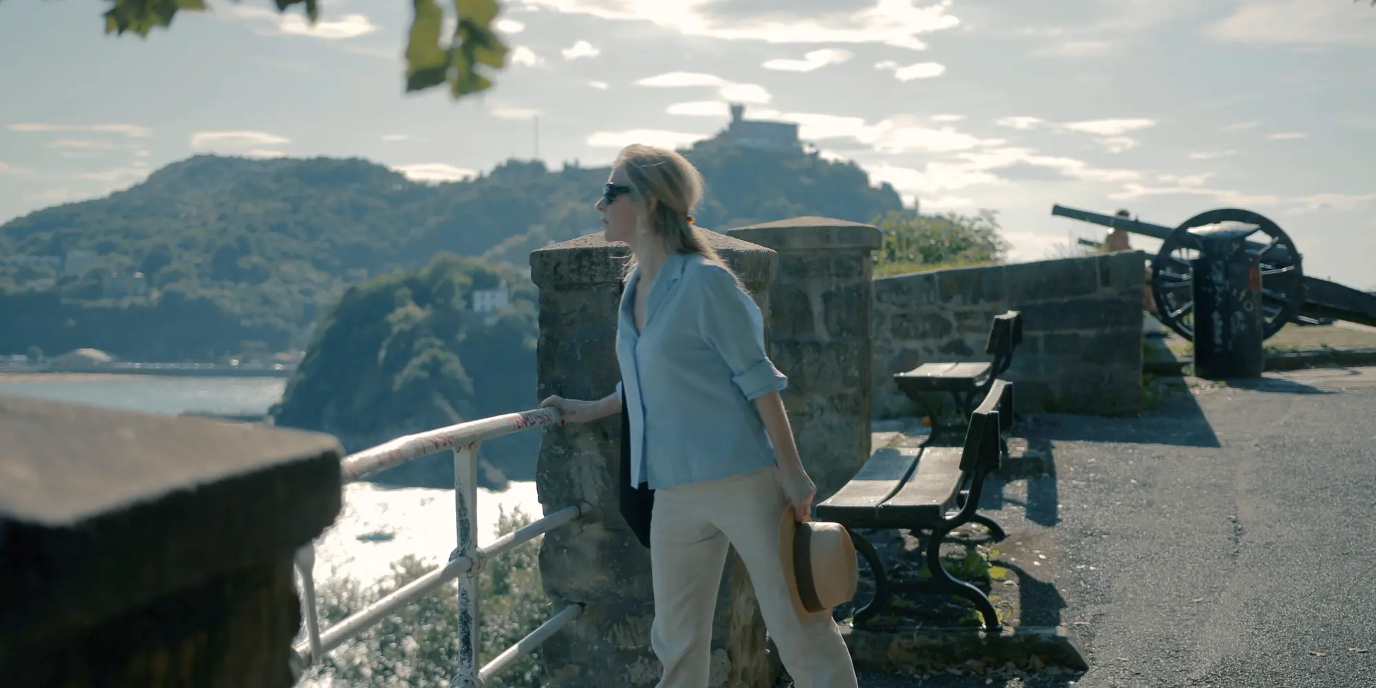 Una mujer rubia con gafas de sol, camisa azul claro y pantalones beige, sostiene un sombrero de paja mientras mira hacia la izquierda, apoyada en una barandilla de piedra y metal en el Monte Urgull. Detrás de ella, se aprecian bancos de madera y un antiguo cañón. Al fondo, se extiende la bahía de La Concha con el mar y el Monte Igueldo, coronado por el Parque de Atracciones y el Funicular. El cielo está parcialmente nublado y el sol ilumina la escena.