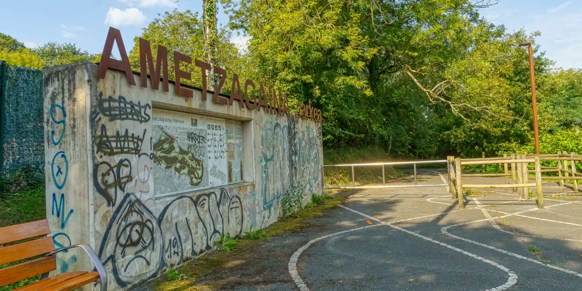 La entrada al "AMETZAGAÑA PARKEA" se muestra con grandes letras metálicas en la parte superior de un muro de hormigón cubierto de grafitis coloridos. En el centro del muro, hay un panel informativo con un mapa y texto sobre el parque. A la izquierda, se ve parte de un banco de madera. A la derecha del muro, hay un camino pavimentado que lleva a una barrera metálica y una valla de madera que marcan la entrada al parque, rodeado de densa vegetación y árboles bajo un cielo parcialmente nublado.