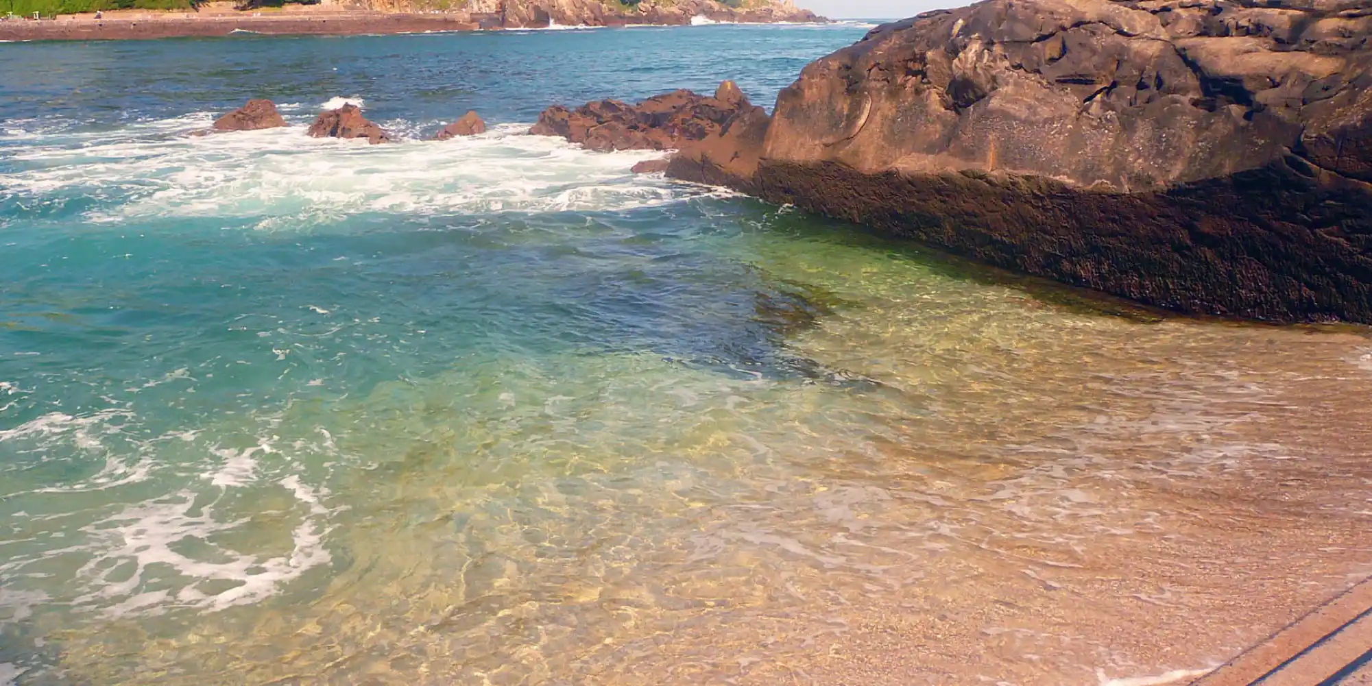Vista de una cala rocosa con aguas cristalinas de tonos verdes y azules, con el sol reflejándose en la orilla de arena clara y rocas oscuras en primer plano y en el fondo.