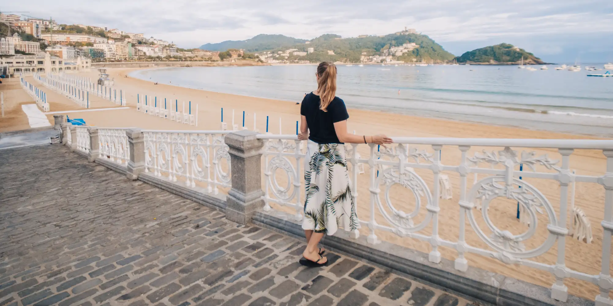 Una mujer de espaldas mirando hacia la playa y la Bahía de La Concha en Donostia-San Sebastián desde el paseo marítimo, con la playa de arena casi vacía y las montañas de la ciudad al fondo.