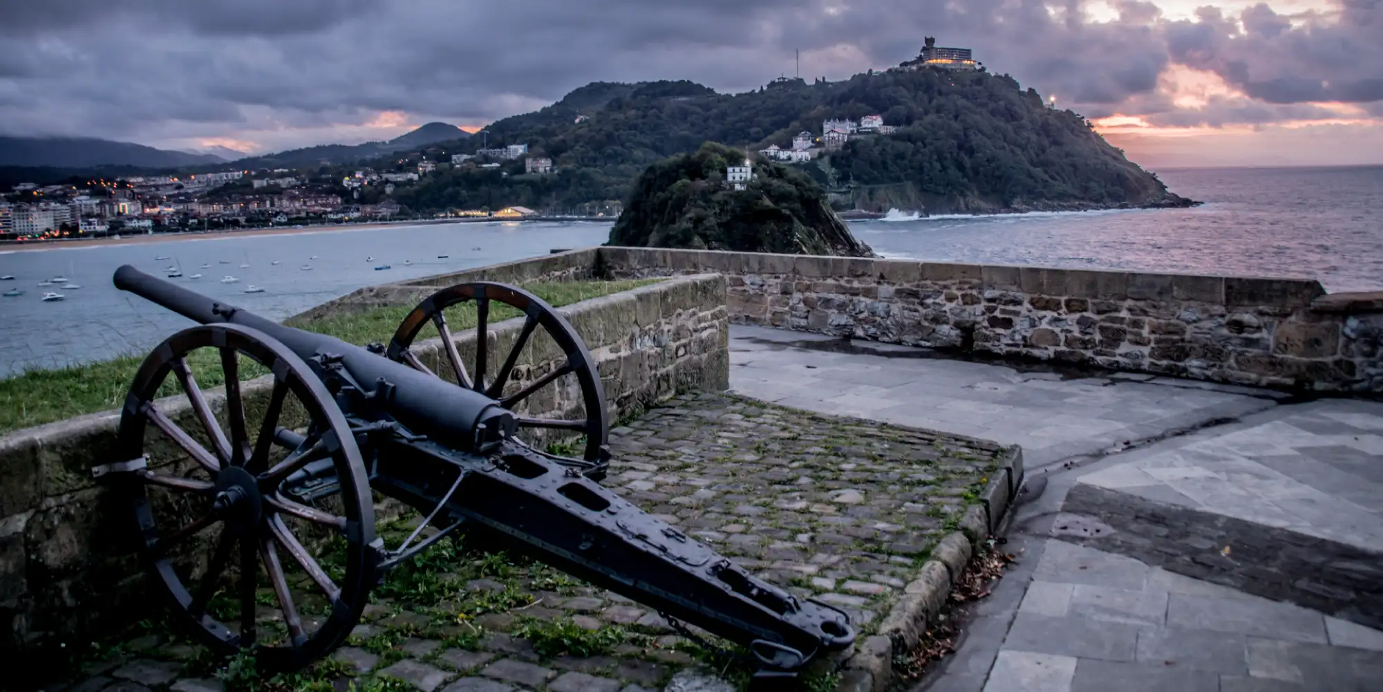 Un antiguo cañón oscuro con ruedas de madera se encuentra en primer plano sobre un empedrado en el Monte Urgull. Detrás del cañón, hay un muro de piedra que ofrece vistas panorámicas de la ciudad de San Sebastián y la bahía de La Concha al anochecer. Las luces de la ciudad comienzan a brillar, y la silueta del Monte Igueldo, con su parque de atracciones iluminado en la cima, es visible al fondo. El cielo presenta tonos de un atardecer nublado, con nubes oscuras y un tenue resplandor anaranjado en el horizonte sobre el mar.