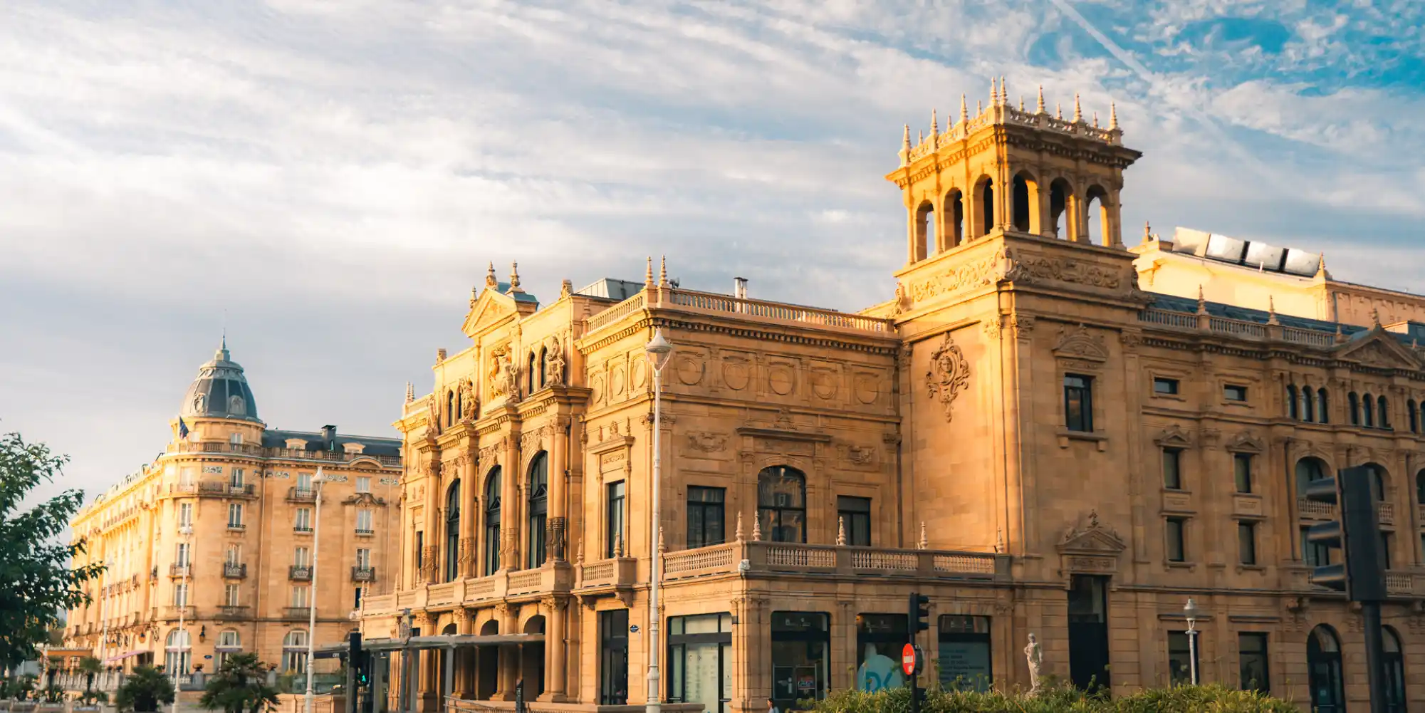 Una gran ópera o teatro de color ocre con rica ornamentación y torres, bajo un cielo parcialmente nublado. A su izquierda, otro edificio clásico con una cúpula.