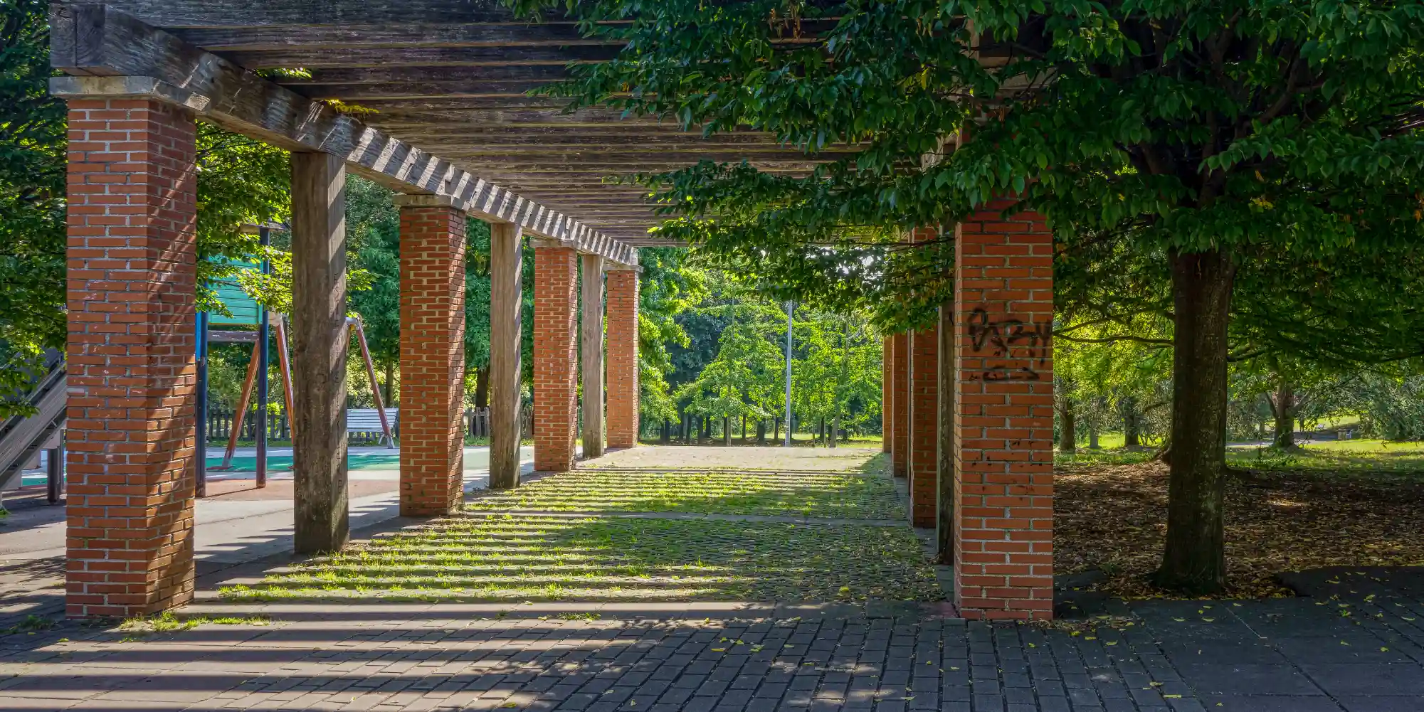 Una larga pérgola con columnas de ladrillo rojizo y una estructura superior de madera oscura se extiende hacia el fondo de la imagen, creando un pasillo sombreado. Rayos de sol se filtran a través de la vegetación que cubre parcialmente la pérgola, proyectando patrones de luz y sombra sobre el suelo adoquinado. A la izquierda, se vislumbra parte de una zona de juegos infantiles con estructuras de colores. Al fondo, se abre una zona verde con árboles exuberantes iluminados por el sol. Una larga pérgola con columnas de ladrillo rojizo y una estructura superior de madera oscura se extiende hacia el fondo de la imagen, creando un pasillo sombreado. Rayos de sol se filtran a través de la vegetación que cubre parcialmente la pérgola, proyectando patrones de luz y sombra sobre el suelo adoquinado. A la izquierda, se vislumbra parte de una zona de juegos infantiles con estructuras de colores. Al fondo, se abre una zona verde con árboles exuberantes iluminados por el sol.