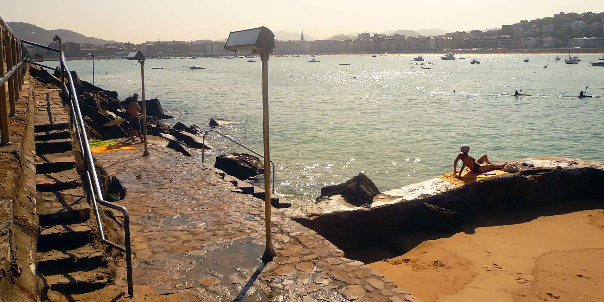 Vista de una playa y la bahía de La Concha en San Sebastián, con una persona tomando el sol en la arena, otra persona saliendo del agua por unas rocas, y varias embarcaciones en el mar bajo un cielo claro.