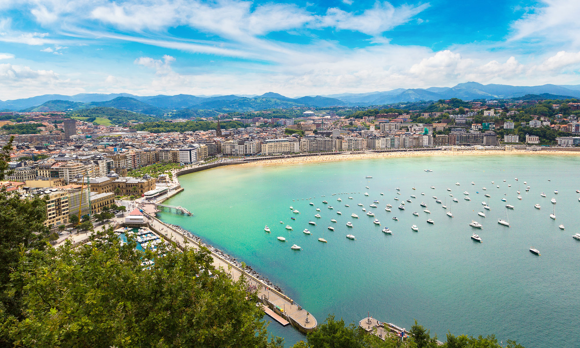 Donostiako Kontxako badiaren ikuspegi panoramikoa, hondartza, itsas ondoko pasealekua eta ur turkesen gainean ainguratutako ontzi ugari ikus daitezke.